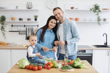 Kahvaltı için yemek pişirirken modern mutfakta poz veren üç çocuklu beyaz bir ailenin portresi. Meraklı kız salata kasesinin yanında dinlenirken gülümseyen anne babalar yemek odasında sarılıyorlar..