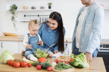 Ürünler ve mutfak gereçleri ile kaplı ahşap masaya yaslanmış kot elbiseli erkek manzarası. Çekici anne salata karışımı olarak kaşık kullanırken aktif çocuk kasedeki yeşillikleri kontrol ediyor..