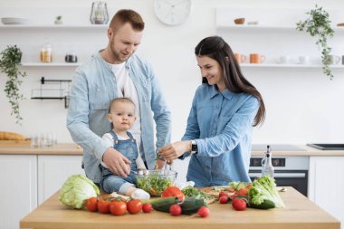 Gülümseyen eşlerin bahçedeki salatayı giydirirken meraklı küçük kız da dairenin mutfağında dinleniyor. Genç, beyaz bir çift ve bebek aile olarak birlikte yemek yapmanın faydalarından faydalanıyor..