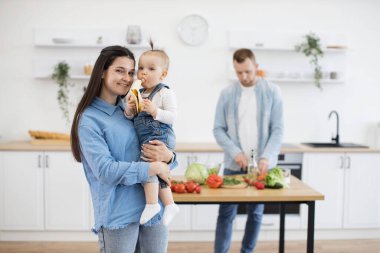 Çekici, uzun saçlı, bebeği elinde tutan ve bebek evde muzla eğlenirken kameraya gülümseyen bir kadın. Anne ve kızı akşam yemeğini beklerken baba sebze salatası yapıyor..