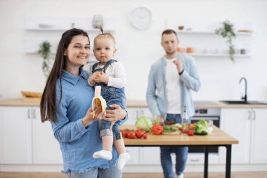 Annelerin ellerinde otururken atkuyruklu, organik muz yiyen küçük sevimli şey. Sorumlu baba, beyaz mutfakta ahşap masada öğle yemeği için sebze kesip pişiriyor..