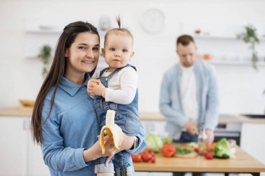 Çekici, koyu saçlı, elinde çocuğu olan anne kameraya bakıp gülümserken ilgili baba da mavi arka planda taze vegan salatası yapıyor. Önemseyen ebeveynler, küçük çocukla birlikte evde dinleniyorlar..