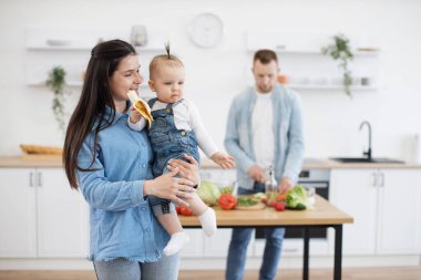 Annelerin ellerinde otururken atkuyruklu, organik muz yiyen küçük sevimli şey. Sorumlu baba, beyaz mutfakta ahşap masada öğle yemeği için sebze kesip pişiriyor..