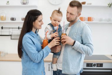 Evli ve çocuklu genç bir çift evdeki rahat mutfakta birlikte mutlu vakit geçiriyorlar. Multivitamin meyve suyu şişesi tutan ve komik suratlı bebeğe bakan neşeli ebeveynler..