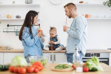 Harika ebeveynler ve çocuklar modern mutfakta yemek yaparken ferahlatıcı bir bardak taze meyve suyunun tadını çıkarıyorlar. Anne ve baba sevimli bir şekilde kızlarına bakıyor. Ailece evde iyi vakit geçirin..