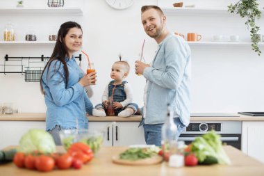 Pazar sabahı mutfakta sağlıklı yemek yapan ve meyve suyu şişeleriyle poz veren aile üçlüsü. Kot pantolon giyen mutlu insanlar kameraya gülümsüyor. Ev eğlencesi ve aşçılık kavramı.