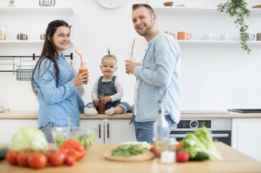 Pazar sabahı mutfakta sağlıklı yemek yapan ve meyve suyu şişeleriyle poz veren aile üçlüsü. Kot pantolon giyen mutlu insanlar kameraya gülümsüyor. Ev eğlencesi ve aşçılık kavramı.