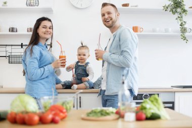 Pazar sabahı mutfakta sağlıklı yemek yapan ve meyve suyu şişeleriyle poz veren aile üçlüsü. Kot pantolon giyen mutlu insanlar kameraya gülümsüyor. Ev eğlencesi ve aşçılık kavramı.