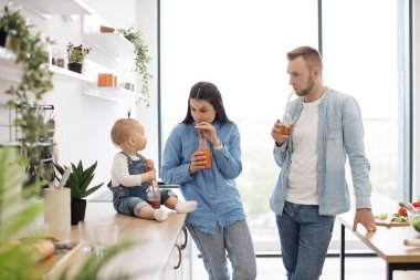 Kendini meyveli organik meyve suyu yudumlamaya adamış ebeveynler mutfaktaki büyük panoramik pencerelerin yanında bebeğe bakıyorlar. Çalışan yüzeyde oturan ve elinde plastik pipet tutan sevimli küçük kız..