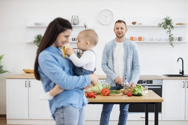 stock image Smiling bearded father cutting vegetables and making healthy dinner while loving mother carrying baby on hands in modern kitchen. Cute toddler treating mommy with tasty banana.