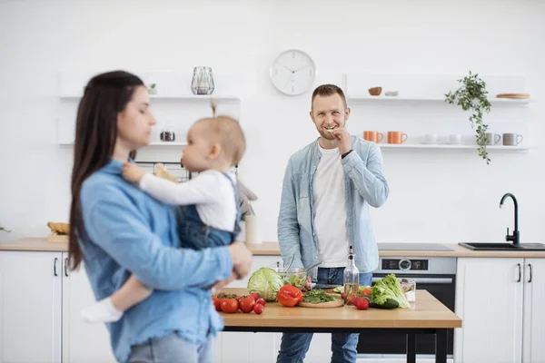 stock image Long-haired brunette mother wearing baby on hands while tall handsome father cooking healthy breakfast for family. Adorable family enjoying spending time together in kitchen. Dieting concept.