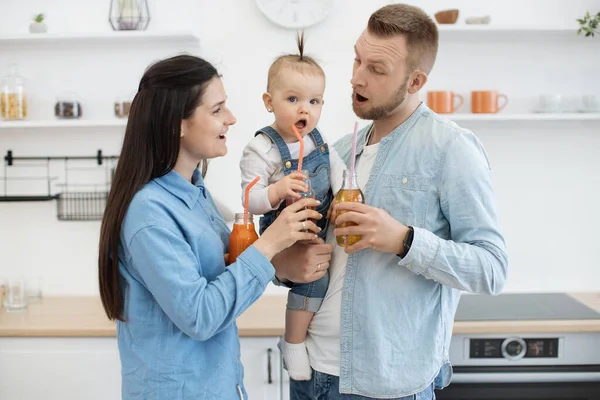 一对年轻夫妇带着孩子在舒适的厨房里共度快乐时光 玩世不恭的父母拿着装有多种维生素汁的瓶子 看着满脸滑稽的婴儿 — 图库照片