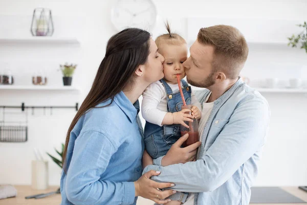 Loving mom and dad in everyday outfit kissing sweet little daughter in cheeks while standing in new modern kitchen. Adorable kid drinking fresh juice through plastic straw. Concept of happy family.