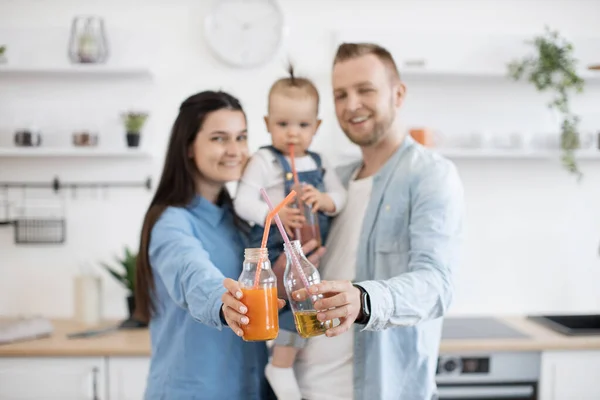 带着爱心的父母牵着小孩的手 在厨房对着相机微笑 模糊的肖像 穿着休闲装 装着新鲜榨果汁装瓶的快乐家庭 — 图库照片