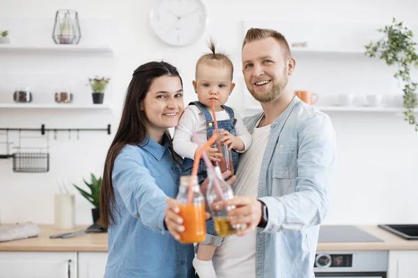 一个快乐的高加索家庭手里拿着装有新鲜果汁的瓶子 对着相机微笑的画像 可爱的父母和带着马尾辫的小宝宝在明亮的厨房里享受美味的饮料 — 图库照片