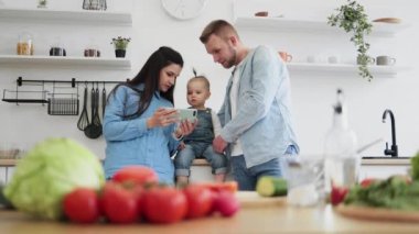 Üç kişilik kot elbiseli mutlu bir aile evdeki mutfak masasının arkasında seyyar ekrana bakıyor. İnternette yemek tarifi arayan ebeveynler ve çocuklar. Ön planda salata için taze malzemeler var..