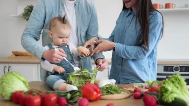 Gülümseyen eşlerin bahçedeki salatayı giydirirken meraklı küçük kız da dairenin mutfağında dinleniyor. Genç, beyaz bir çift ve bebek aile olarak birlikte yemek yapmanın faydalarından faydalanıyor..