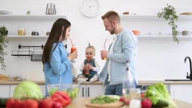 Harika ebeveynler ve çocuklar modern mutfakta yemek yaparken ferahlatıcı bir bardak taze meyve suyunun tadını çıkarıyorlar. Anne ve baba sevimli bir şekilde kızlarına bakıyor. Ailece evde iyi vakit geçirin..