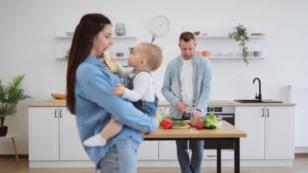 在现代厨房里 满脸胡须的父亲一边切蔬菜一边做着健康的晚餐 一边爱着手里抱着孩子的母亲 可爱的幼儿用美味的香蕉喂妈妈 — 图库视频影像