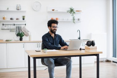 Gözlüklü neşeli sakallı adam, geniş yemek odasında masa başında otururken modern laptop kullanıyor. Gülümseyen Hintli serbest yazar, çalışma saatlerini evde geçirirken esnek program kullanıyor..