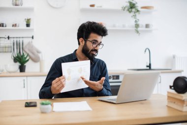 Çekici Hintli erkek, evdeki bilgisayar aracılığıyla çevrimiçi toplantı sırasında konuşurken elinde çizelgelerle kâğıt tutuyor. Nitelikli veri analisti mutfak içindeki iş büyümesi hakkında bilgi veriyor.