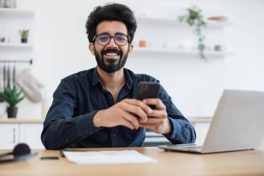Evde modern işyerinde kalırken elinde akıllı telefon tutan gözlüklü pozitif Hintli erkek. Verimli serbest çalışan, kapalı alanda işverenin projesini yaparken meslektaşına mesaj gönderiyor..