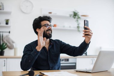 Gülümseyen sakallı yetişkin gözlüklü. Mutfaktaki ofis masasında akıllı telefonu kaldırırken selam veriyor. Neşeli Hintli iş adamı, çalışma saatleri boyunca katılımcıları selamlıyor..