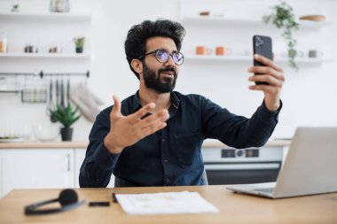 Gözlüklü çekici genç adam mutfak arka planında web kamerasıyla konuşurken telefonu tutuyor. Yakalı tişörtlü mutlu Hintli ev içindeki modern cihazlar aracılığıyla online konferans düzenliyor..