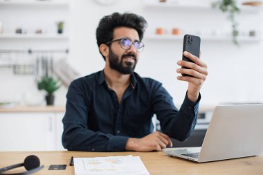 Gözlüklü çekici genç adam mutfak arka planında web kamerasıyla konuşurken telefonu tutuyor. Yakalı tişörtlü mutlu Hintli ev içindeki modern cihazlar aracılığıyla online konferans düzenliyor..