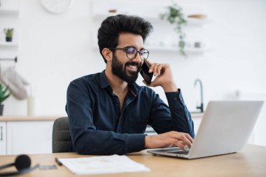 Geniş yemek odasında taşınabilir bilgisayar kullanırken akıllı telefondan konuşan sıradan giyinmiş neşeli genç adam. Verimli Hintli serbest yazar, uzak bir şirketin müşterisiyle iş görüşmesi yapıyor..