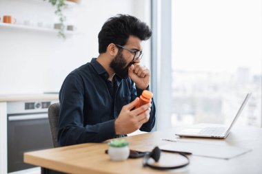 Dairenin panoramik penceresinin yanında ofis masasında otururken öksüren gözlüklü çekici Hintli erkek. Verimli bir işletme sahibi, evde hastalıkla savaşırken şirketi yönetiyor..