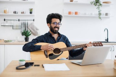 Geniş apartmandaki taşınabilir bilgisayar webcam 'ine gülümserken telli çalgılar tutan neşeli bir Arap. Mutlu gözlüklü erkek modern teknolojileri kullanarak online gitar dersi veriyor..