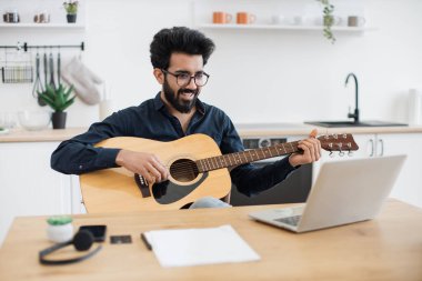 Geniş apartmandaki taşınabilir bilgisayar webcam 'ine gülümserken telli çalgılar tutan neşeli bir Arap. Mutlu gözlüklü erkek modern teknolojileri kullanarak online gitar dersi veriyor..