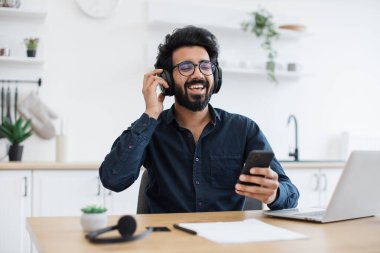 Koyu renk gömlek ve gözlük takan tutkulu genç adam yemek odasında dinlenirken kulaklıkla yüksek sesli müzik dinliyor. İçeriden indirme yapmak için cep telefonu uygulaması kullanan mutlu Arap yetişkin.