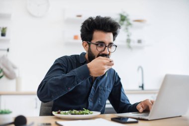 Mutfaktaki ahşap masada portatif bilgisayarda yazarken gözlük takıp sebze yiyen etkili bir Hintli işçi. Uzaktan bağımsız çalışan öğle yemeği molasında vakit kaybetmeden iş yapıyor..