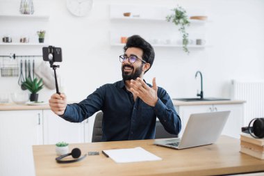 Gözlüklü neşeli Arap yetişkin dairede yazı masasında çalışırken elinde akıllı telefonuyla tripodla geziyor. Gülümseyen yönetici mobil uygulama kullanılarak başlatılan projeyle ilgili video raporunu kaydediyor.