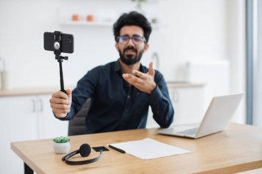 Gözlüklü yorgun Hintli adamın dizüstü bilgisayarla masada otururken tripoda yerleştirdiği modern cep telefonuna odaklan. Uzman danışman uzak işyerinden canlı yayın detaylı raporu.