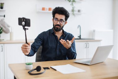 Gözlüklü yorgun Hintli adamın dizüstü bilgisayarla masada otururken tripoda yerleştirdiği modern cep telefonuna odaklan. Uzman danışman uzak işyerinden canlı yayın detaylı raporu.