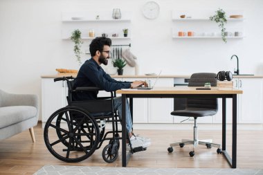 Tekerlekli sandalyedeki Hintli bir erkeğin modern dairenin açık-planlı mutfağında yazı masasında çalışmasının yan görüntüsü. Özürlü genç yetişkinler işe evde e-posta hesabıyla başlıyorlar..