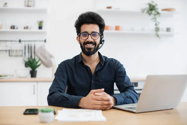 stock image Portrait of smiling person wearing headset with microphone posing with documents and portable computer. Cheerful arabian consultant offering guidance to clients over digital platform from home.