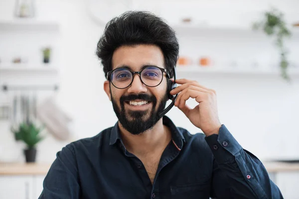 Close up view of indian man holding microphone of hands-free headset while smiling at camera on kitchen background. Happy professional working virtually as staff member for fully remote company.