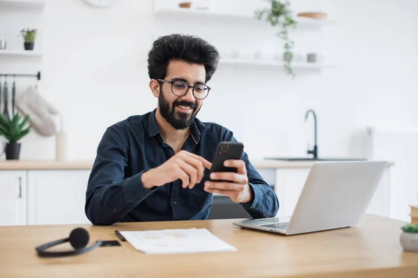 現代の職場で自宅にいる間 スマートフォンを持って眼鏡をかけて積極的なインドの男性 屋内で雇用主のためのプロジェクトを実行しながら 同僚に効率的なフリーランスの労働者のテキストメッセージ — ストック写真