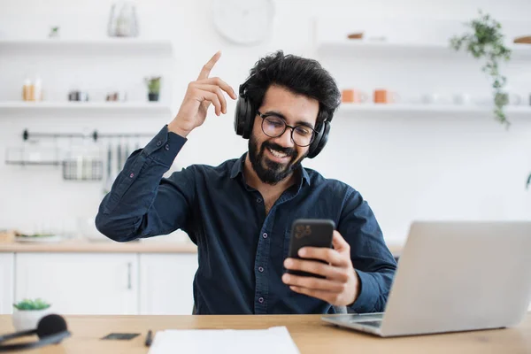 Joyous man in glasses and button-down shirt raising index finger while looking at smartphone in kitchen. Cheerful indian adult singing along while hearing loud tunes in headphones during break.