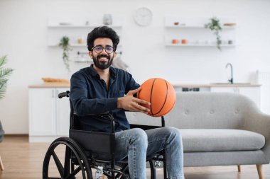 Tekerlekli sandalyedeki çekici Arap erkek portresi daire mutfağında basketbolla poz veriyor. Gülümseyen sakallı insan içeride fiziksel aktivitenin gerçek sağlığına kavuşuyor..