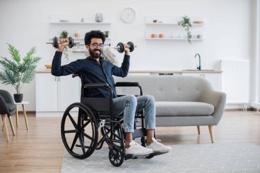 Tekerlekli sandalyede ağırlık kaldıran yakışıklı Hintli adamın kapalı alanda hareket kabiliyeti sınırlıyken tam boy görüntüsü. Mutfakta çalışırken orta yoğunluklu aktivite kullanarak gülümseyen bir yetişkin..