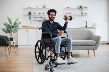 Geniş açılı, hareketlilik bozukluğu olan genç Hintli erkek açık-plan mutfakta ağırlık egzersizi yapıyor. Eğitimden sağlık yardımı alan kot pantolonlu, gözlüklü bir yetişkin..