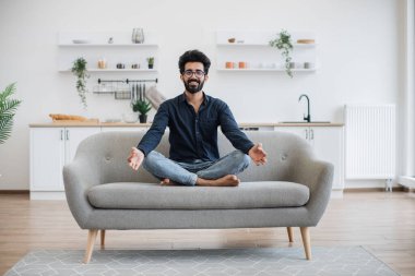 Modern apartmandaki açık-plan mutfağı olan bir kanepede bağdaş kurmuş, gündelik giysiler içinde gülümseyen genç insan. Rahat Hintli erkek, güneşli bir günde parmaklarıyla Gyan Mudra 'da meditasyon yapıyor..
