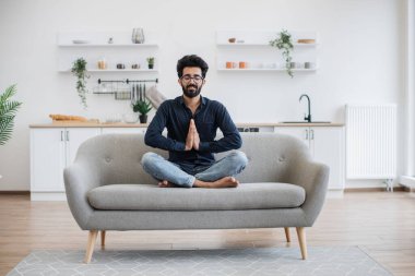 Kotlu ve koyu renk gömlekli genç sakallı adam yoga pozunda kanepede uzanırken Anjali Mudra 'da el ele tutuşuyor. Evde bağdaş kurarak otururken stresi azaltan çekici Hintli yetişkin..