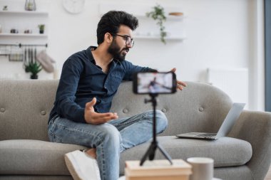 Şık saç kesimi olan Hintli adam sosyal ağda canlı yayında modern laptoptan bilgi okuyor. Çevrimiçi eğitim için tripodlu modern akıllı telefon kullanılması talebi.