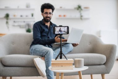 Kanepede dizüstü bilgisayarla oturan ve akıllı telefon kamerasıyla konuşan neşeli bir Arap 'ın bulanık arka planı. Sosyal medyası için olumlu erkek blogcu video kaydı.